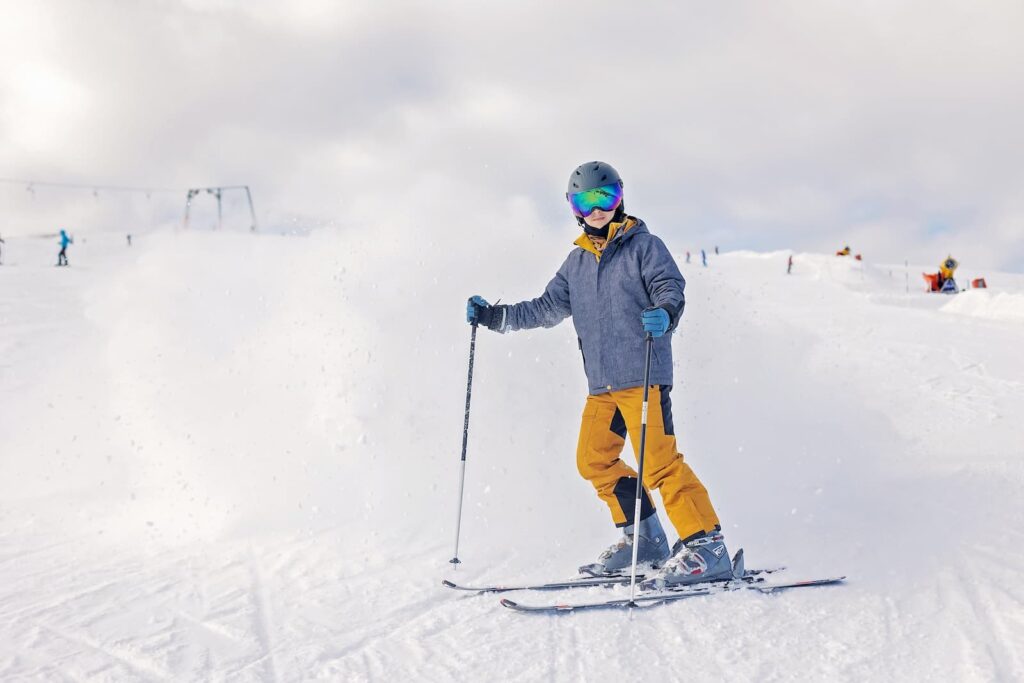 A photo shows a young person skiing on a snowy mountain. They are wearing a gray jacket, bright yellow pants, a black helmet, and reflective goggles that have a rainbow tint. They are holding two ski poles and are looking directly at the camera with a slight smile. The background is a white, snowy slope with other skiers and ski lifts in the distance, under a cloudy white sky.
