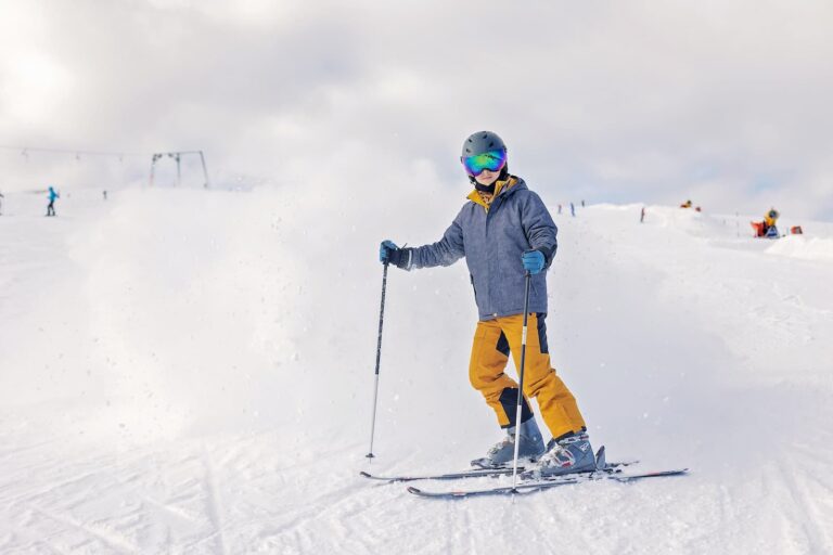 A photo shows a young person skiing on a snowy mountain. They are wearing a gray jacket, bright yellow pants, a black helmet, and reflective goggles that have a rainbow tint. They are holding two ski poles and are looking directly at the camera with a slight smile. The background is a white, snowy slope with other skiers and ski lifts in the distance, under a cloudy white sky.